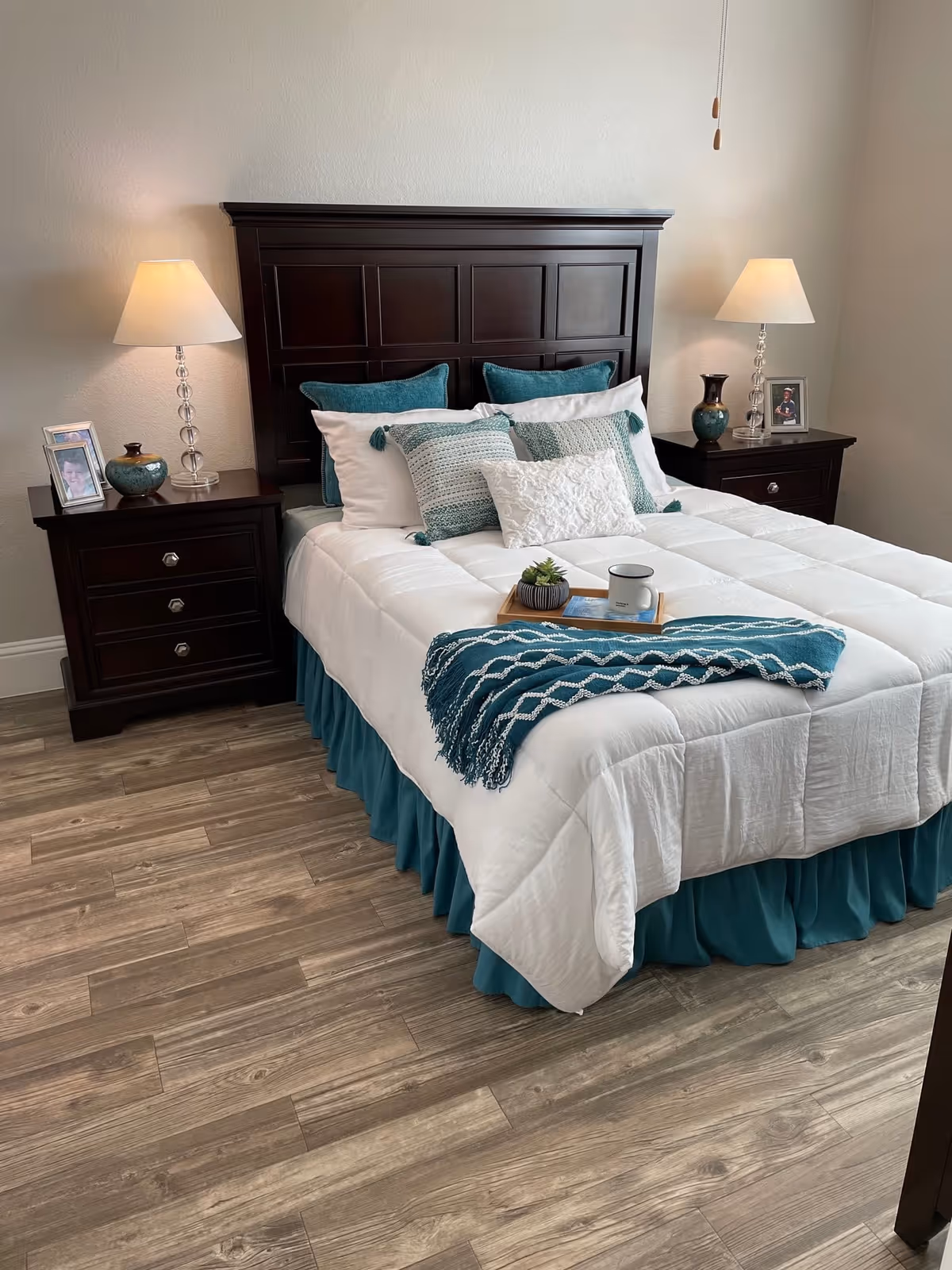 A neatly made bed with a dark wooden headboard and white bedding accented with teal pillows and a teal throw blanket. There are two matching dark wooden nightstands on either side of the bed, each with a lamp, decorative items, and framed photos. The room has wood flooring and light-colored walls.