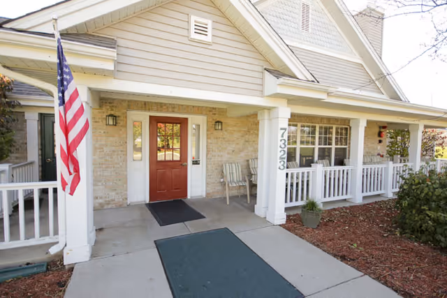 Front exterior view of a residential-style building with a covered porch, a red door, two chairs, an American flag mounted on a white column, and the number 7323 displayed on the column.
