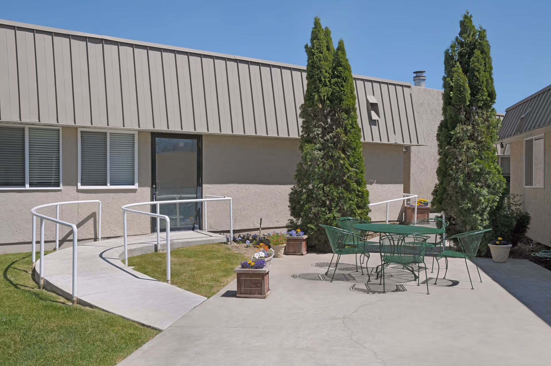 Sunlit courtyard with a concrete wheelchair ramp, potted flowers, evergreen trees and a green metal table and chairs beside a beige building.