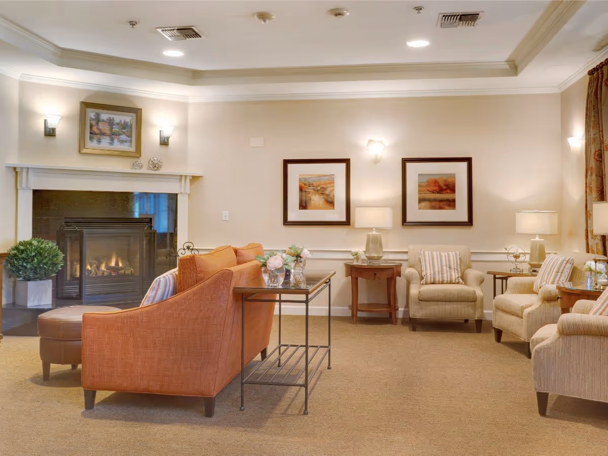 Cozy furnished lounge with a lit fireplace, an orange sofa, several armchairs, side tables, lamps, and framed artwork.