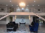 Spacious modern living room area with two black sofas and two blue armchairs arranged around a glass coffee table on a patterned rug. The room features a large chandelier hanging from the ceiling, a white staircase with metal railings on both sides leading to an upper level, and multiple recessed ceiling lights.