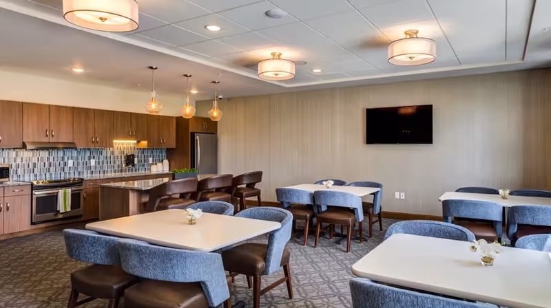 A modern dining area in a senior living facility featuring several square tables with blue upholstered chairs. The room includes a kitchen area with wooden cabinets, a stainless steel refrigerator, stove, and a kitchen island with bar stools. The walls are light-colored, and a flat-screen TV is mounted on one wall. Ceiling lights provide warm illumination.