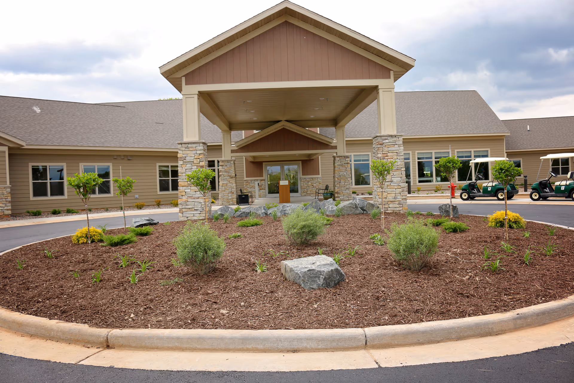 Front exterior view of Dove Healthcare - Rice Lake building with a covered entrance supported by stone pillars, a circular landscaped area with small trees and shrubs, and golf carts parked on the right side.