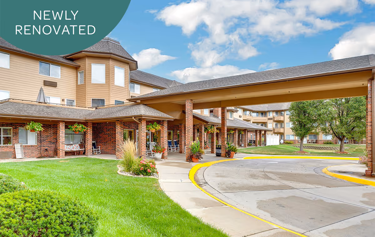 Covered entrance and circular drive of a newly renovated multi-story senior living building with brick facades, hanging plants, and landscaped lawns.
