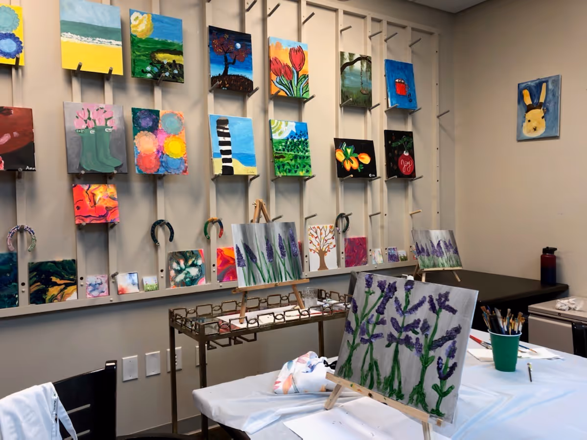 A room with multiple colorful paintings displayed on a wall-mounted rack, featuring various subjects like flowers, landscapes, and abstract designs. In the foreground, there are easels holding paintings of purple flowers on a table covered with a white cloth. Paintbrushes and art supplies are also visible on the table.