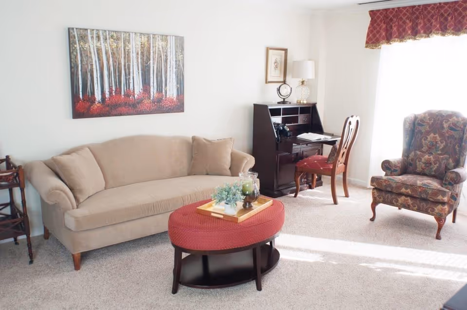 A bright living room with a beige sofa, a red oval ottoman with a wooden tray holding decorative items, a dark wooden desk with a chair, a floral upholstered armchair, and a large window with a red valance allowing sunlight to fill the room. A painting of birch trees with red foliage hangs on the wall above the sofa.