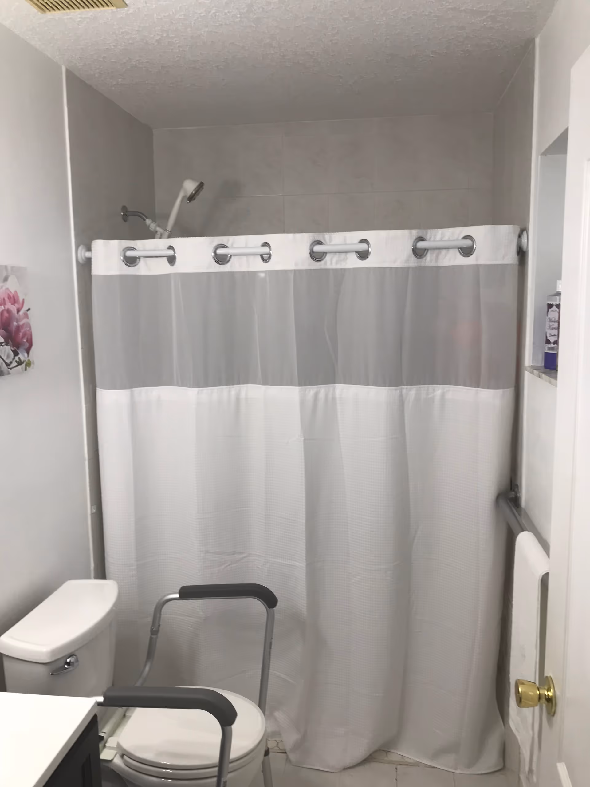 Small bathroom with toilet, shower curtain, grab bars, and a shower chair.