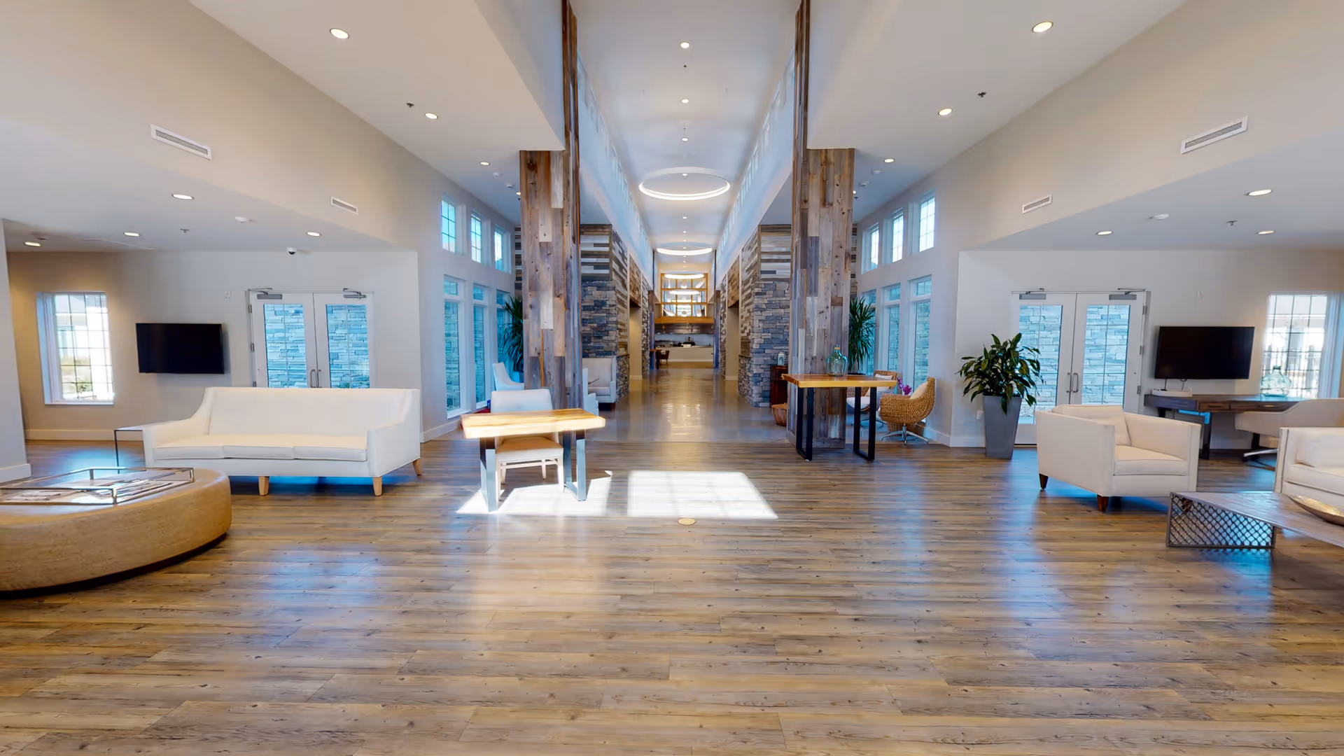 Spacious and well-lit common area in a senior living facility with wooden flooring, white sofas, armchairs, small tables, large windows, and modern ceiling lights. The room has a high ceiling with wooden beams and stone pillars along the sides.