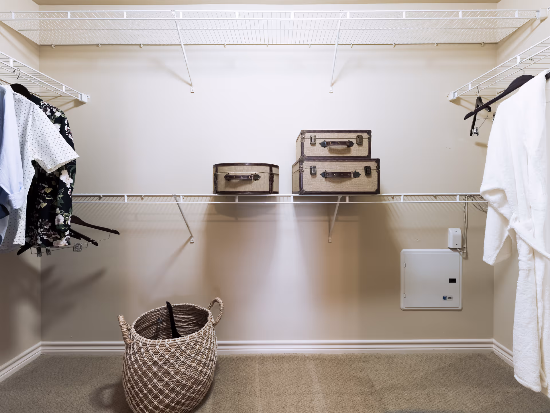 A spacious walk-in closet with beige walls and carpeted floor. The closet has white wire shelving on three sides. On the left side, several shirts are hanging on hangers. On the right side, a white bathrobe is hanging. On the middle shelf, there are three vintage-style suitcases stacked. On the floor, there is a woven basket with handles.