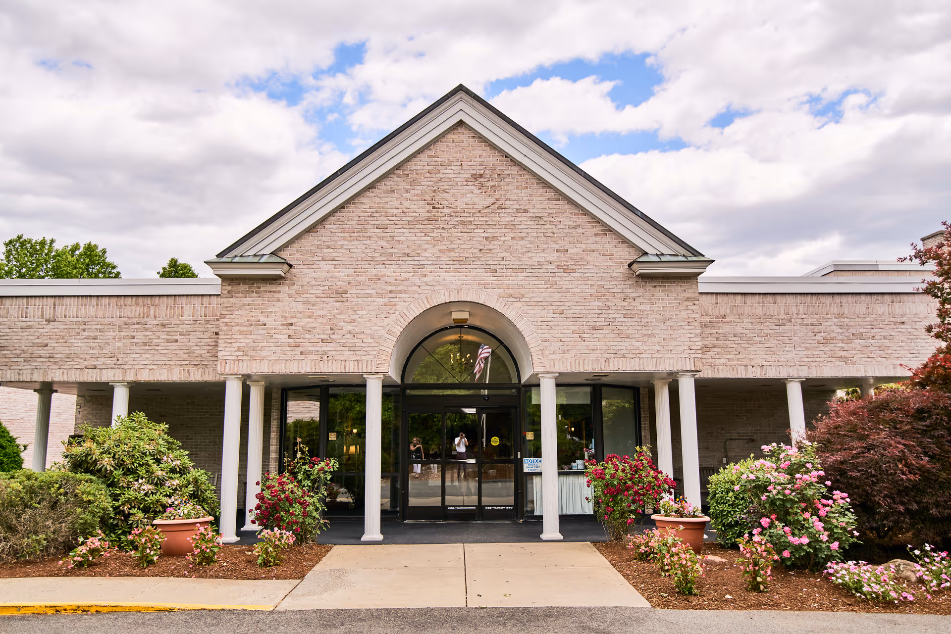 Front exterior view of a brick building with a peaked roof and white columns at the entrance. There are glass double doors under an archway, with flower pots and bushes on either side of the walkway leading to the entrance. The sky is partly cloudy.