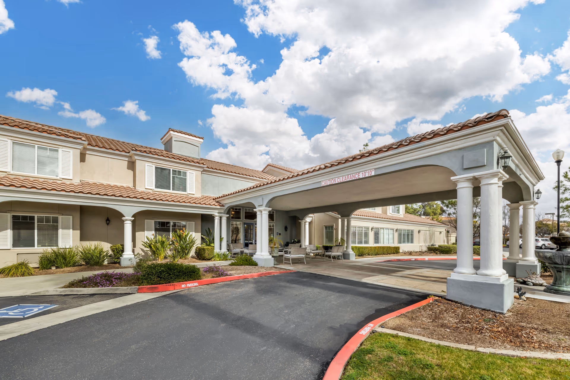 Exterior view of Brookdale Murrieta senior living facility showing the entrance with a covered driveway supported by white columns, beige building with tiled roof, windows with white shutters, landscaped bushes and plants, and a partly cloudy sky.