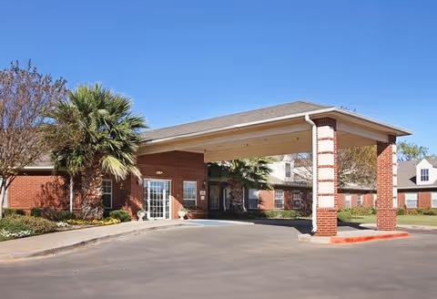 Exterior view of a single-story brick building with a covered entrance supported by brick columns. There are palm trees and other landscaping around the building, and a paved driveway leading to the entrance.