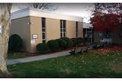 Front exterior of a low brick health and rehabilitation center with a sidewalk, trimmed shrubs, benches, and a tree.