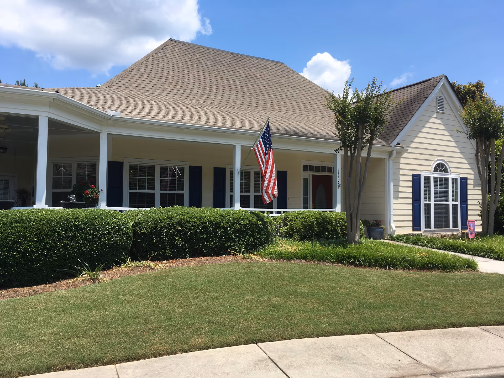 Exterior view of a single-story building with beige siding and a large front porch. The porch has white columns and an American flag hanging from one of them. There are neatly trimmed bushes and a well-maintained lawn in front of the building. The sky is partly cloudy with blue patches visible.