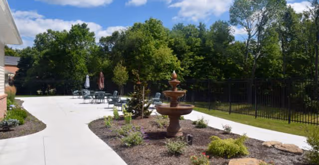 Sunny outdoor courtyard with a central tiered fountain, paved walkways, patio tables with umbrellas, and trees and fencing in the background.