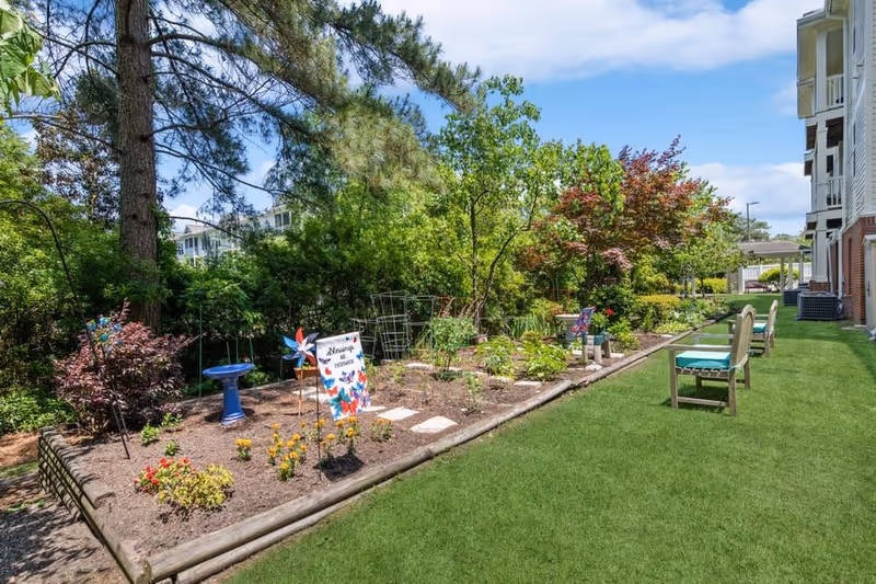 A well-maintained outdoor garden area with various plants and flowers, a birdbath, colorful pinwheels, and a sign that reads 'Blessings for Veterans'. There are several chairs with cushions placed on the grass beside the garden, and a multi-story building is visible on the right side under a partly cloudy sky.