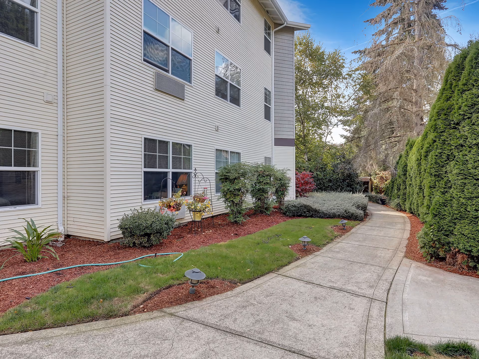 A paved walkway curves alongside a beige multi-story building with several windows. The walkway is bordered by well-maintained landscaping including green grass, shrubs, small trees, and mulch beds. Tall evergreen trees line the right side of the path, and the sky is partly cloudy.