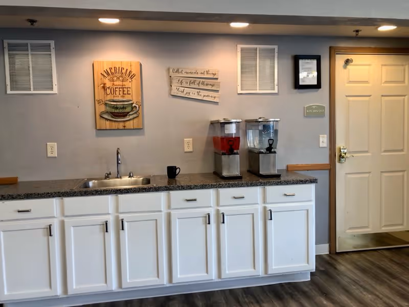 Interior view of a kitchenette area with white cabinets and a granite countertop. There is a small sink with a faucet, a black coffee mug, and two beverage dispensers containing red and clear liquids. On the wall above the countertop, there are two decorative signs, one featuring a coffee cup and the other with an inspirational quote. To the right, there is a closed white door with a gold-colored handle and a small sign that reads 'KITCHEN OUT'. The floor has wood-like vinyl planks.