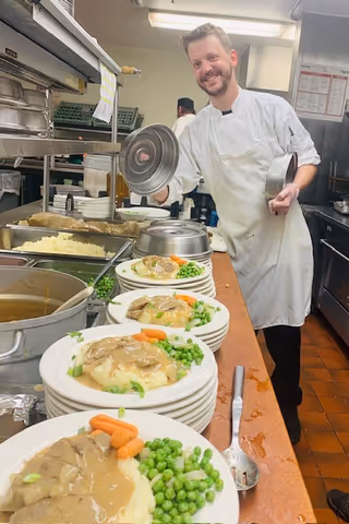 A chef in a white apron and uniform is smiling while plating meals in a commercial kitchen. Several plates with mashed potatoes, gravy, peas, and carrots are lined up on a counter. Kitchen equipment and food trays are visible in the background.