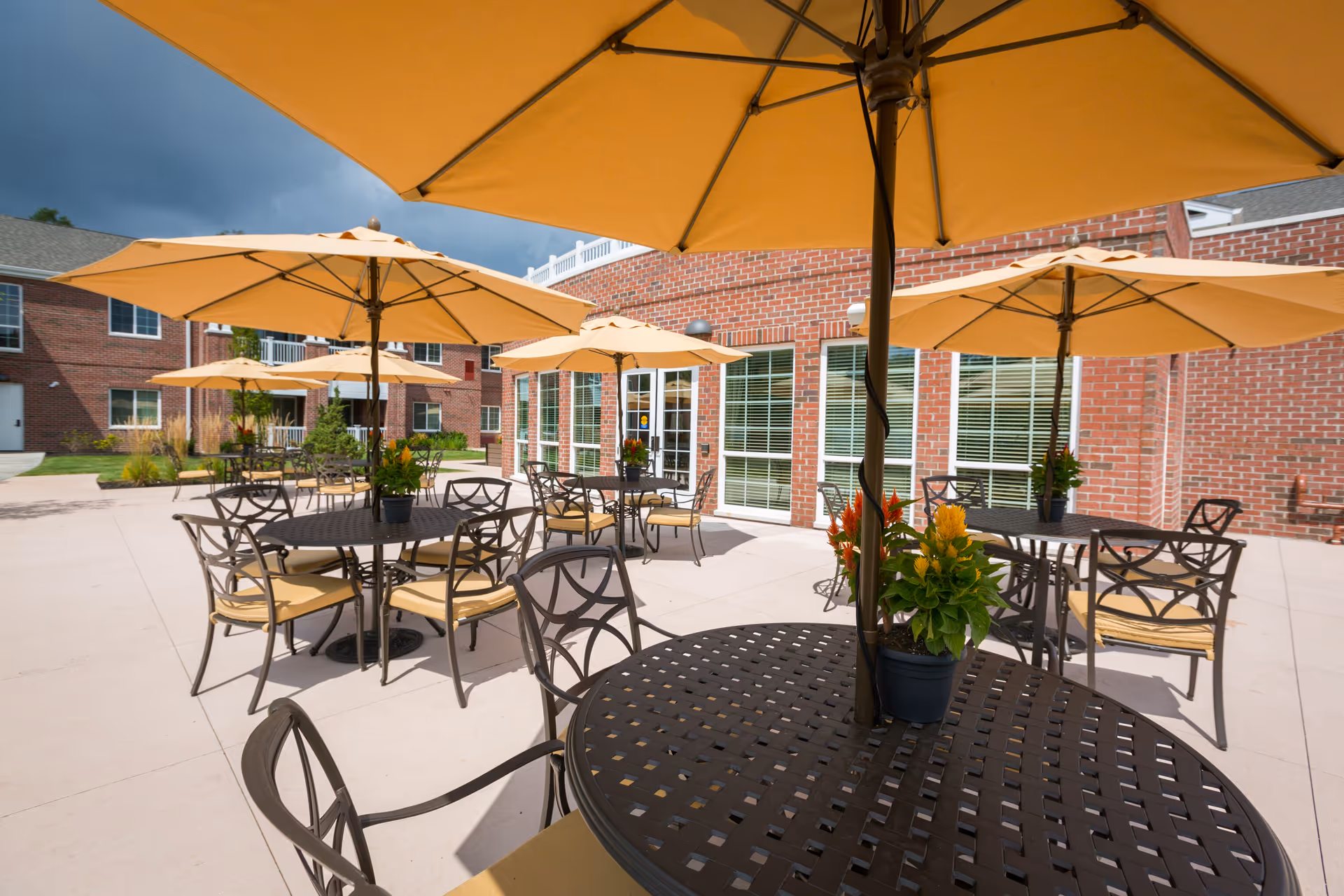Outdoor patio area with multiple round metal tables and chairs, each table shaded by a large yellow umbrella. Potted plants with colorful flowers are placed on the tables. The patio is adjacent to a red brick building with large windows and a door.