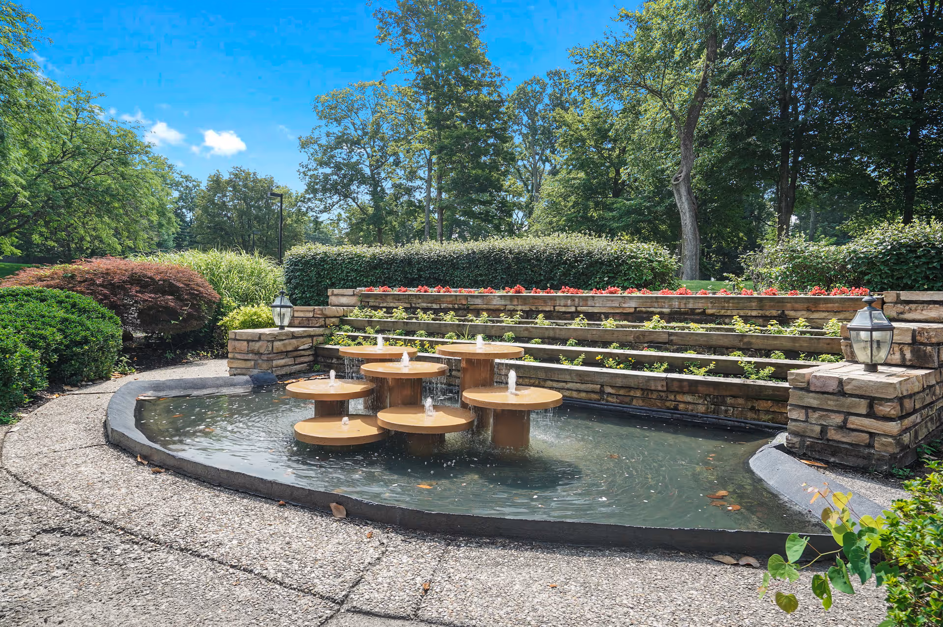 Outdoor garden area with a circular water fountain featuring multiple small water jets. The fountain is surrounded by a curved stone pathway and tiered stone planters filled with green plants and red flowers. Trees and bushes provide a lush green backdrop under a clear blue sky.