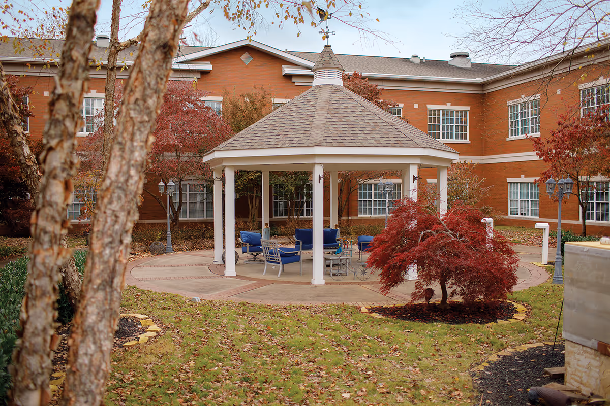 Outdoor gazebo with a shingled roof and white pillars surrounded by a garden with trees and shrubs showing autumn colors. Blue cushioned chairs and a table are arranged under the gazebo. A brick building with multiple windows is visible in the background.