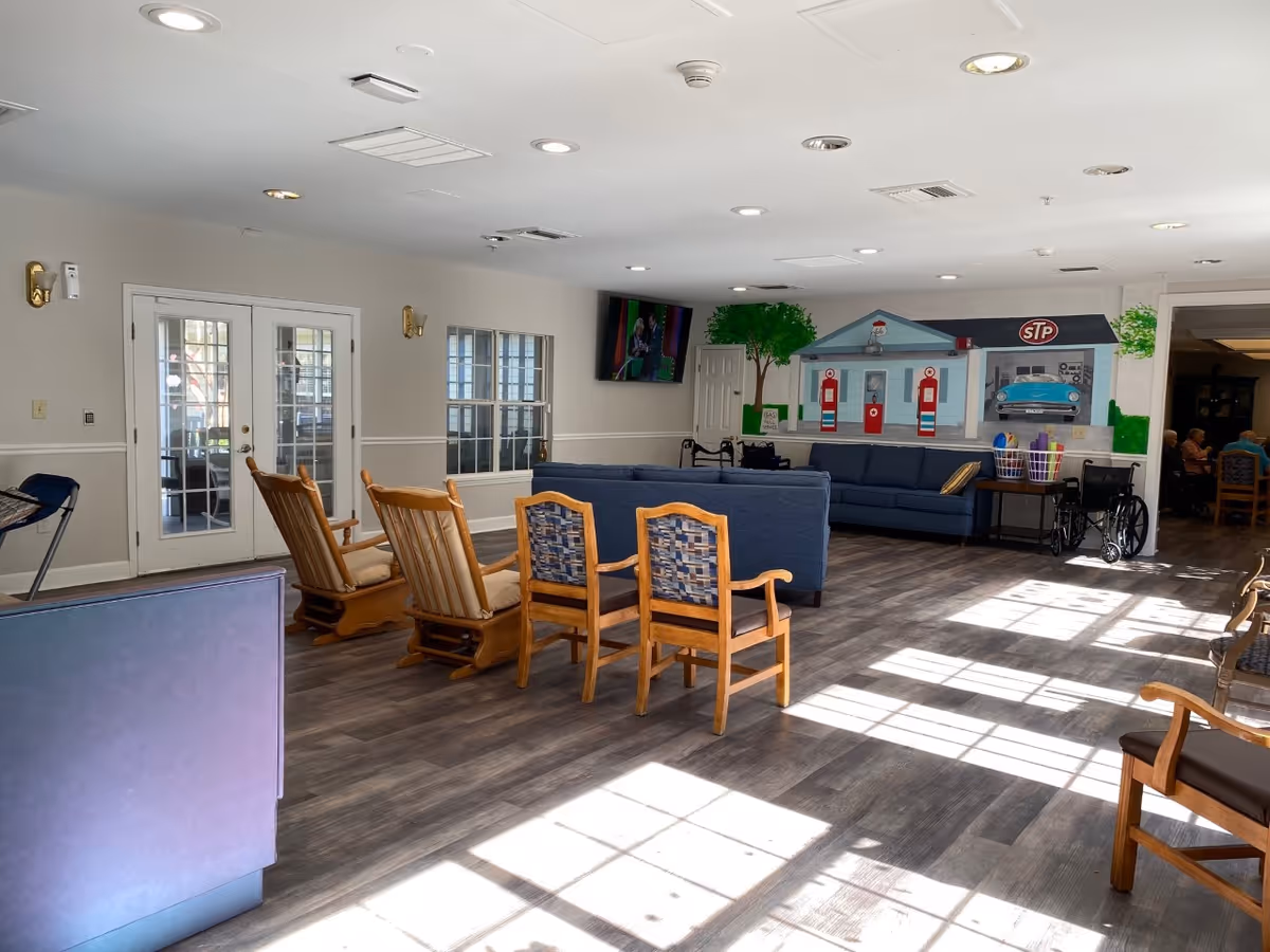 A spacious common area in a senior living facility with wooden rocking chairs and cushioned chairs arranged around blue sofas. There is a mural on the far wall depicting a vintage gas station with a blue car and gas pumps. A TV is mounted on the wall above the sofas, and sunlight streams in through windows, casting shadows on the wooden floor. A wheelchair and a basket with rolled-up mats are visible near the sofas. French doors and windows are on the left wall.