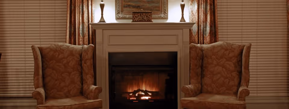 Two upholstered wingback chairs flank a lit fireplace with a mantel, lamps, and window blinds.