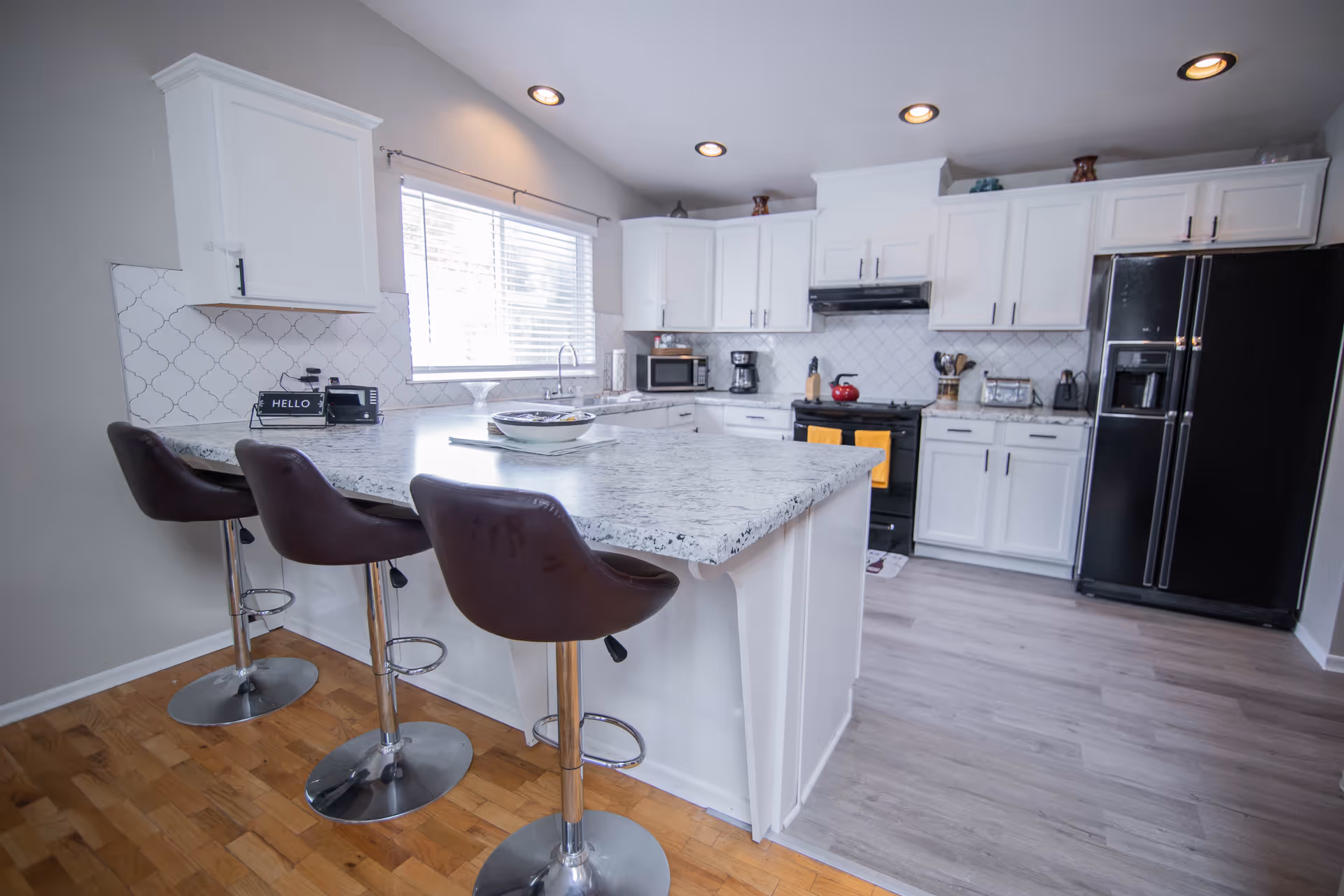 Bright modern kitchen with a marble island, three bar stools, white cabinets, and black appliances.