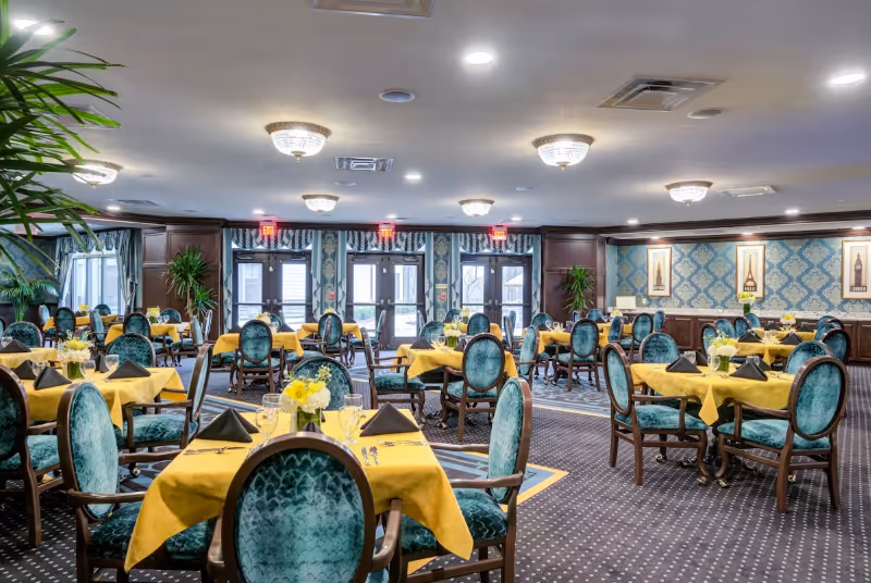 A spacious dining room with multiple tables covered in yellow tablecloths and set with black napkins, glassware, and floral centerpieces. The chairs have teal upholstery and wooden frames. The room has patterned carpet, decorative wallpaper, framed artwork on the walls, and several ceiling light fixtures. Large windows and glass doors are visible at the far end, allowing natural light to enter.