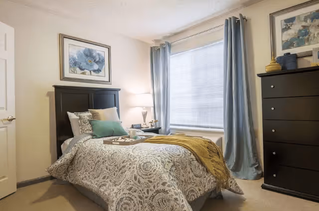 A cozy bedroom with a single bed covered in a patterned comforter and multiple pillows. A wooden headboard is behind the bed, and a tray with a cup and a book is placed on the bed. To the right, there is a tall dark wooden dresser with decorative items on top. A large window with closed blinds and light blue curtains is on the far wall. A framed floral artwork hangs above the headboard and another above the dresser. A bedside table with a lamp and a clock is next to the bed.