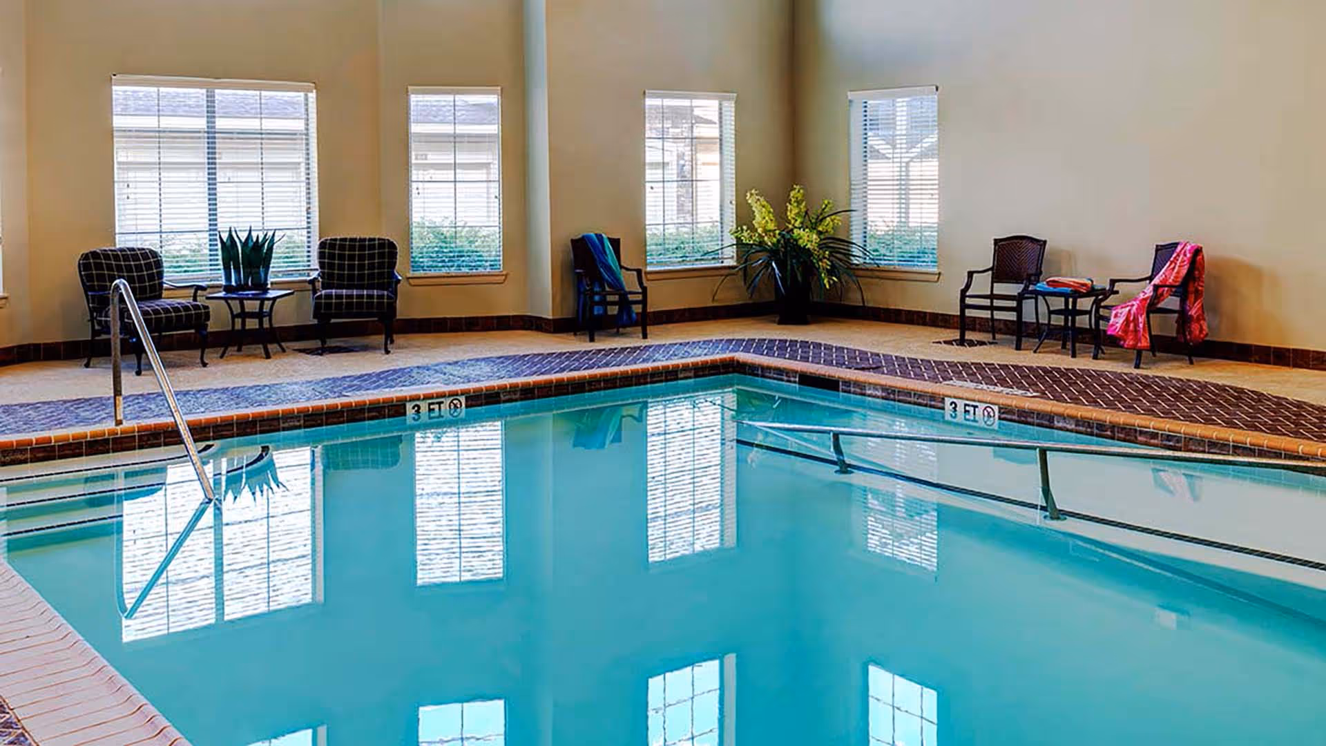 Indoor swimming pool with clear blue water surrounded by beige tiled floor and brick trim. Several chairs and small tables with plants and towels are arranged around the pool area. Large windows with blinds allow natural light to enter the room.