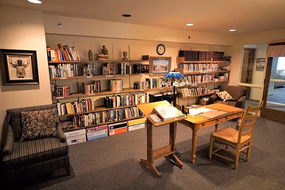 A cozy reading room in a senior living facility featuring a wooden desk with a Tiffany-style lamp, an open book stand, a wooden chair, two armchairs with patterned cushions, and multiple shelves filled with books. The room has beige walls, carpeted floor, and framed artwork on the walls.