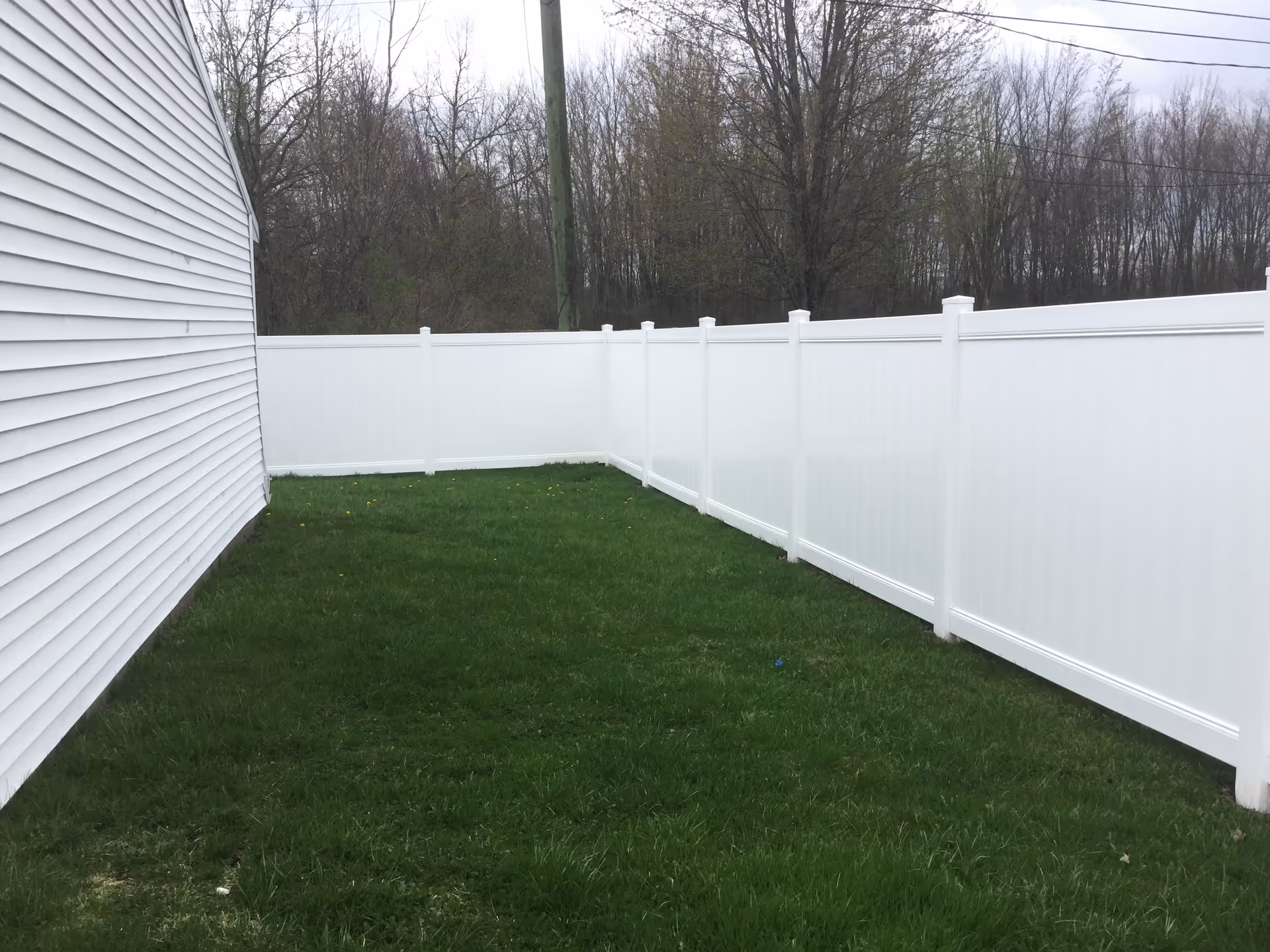 Narrow grassy backyard bordered by a white vinyl privacy fence and the side of a white house with trees beyond.