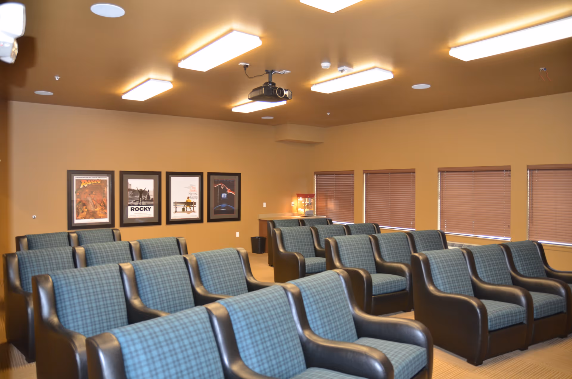 Small movie screening room with rows of upholstered armchairs, a ceiling projector, movie posters on the wall and a popcorn machine.
