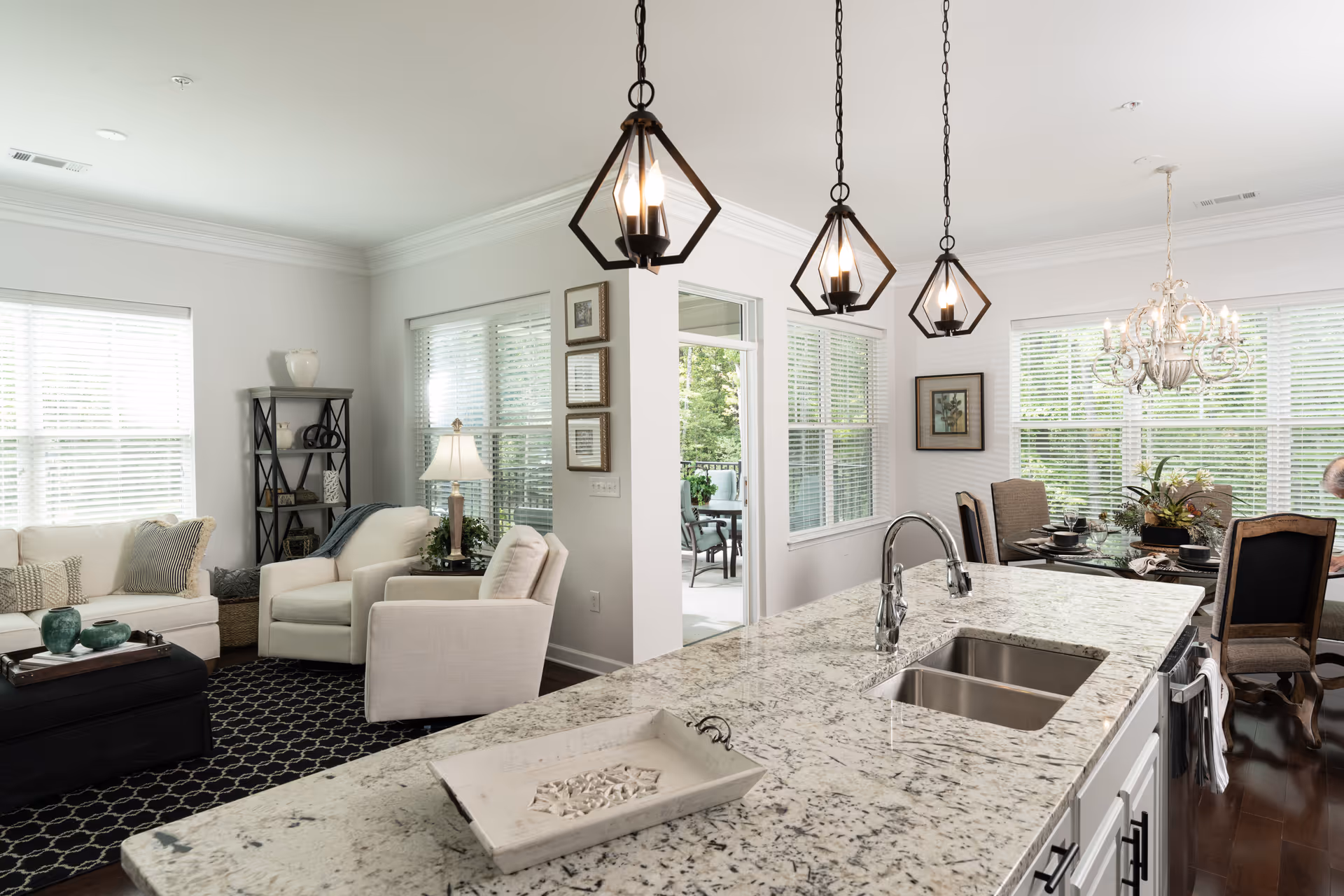 Bright and modern open-concept living space featuring a kitchen island with a granite countertop and dual sinks, three geometric pendant lights hanging above, a cozy living room area with white sofas and armchairs, a black patterned rug, a black shelving unit, and a dining area with a chandelier and a table set for four near large windows with white blinds.