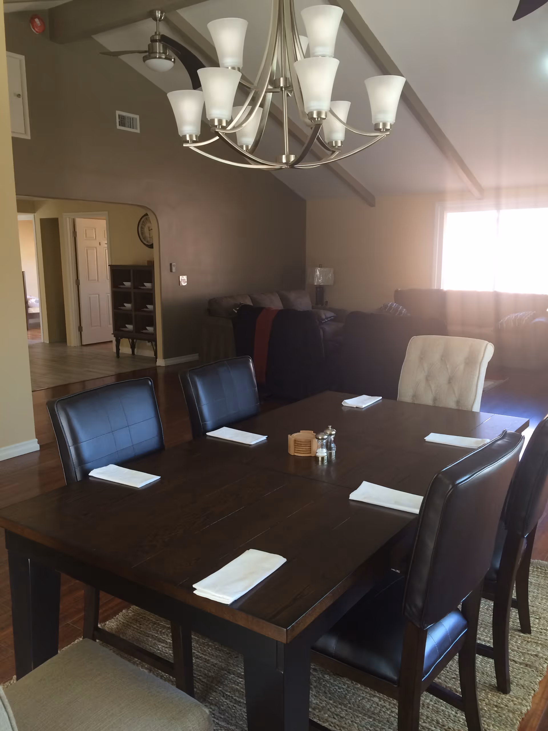 A dining area with a dark wooden table set for six with white napkins on each place. The chairs are a mix of black leather and beige fabric. Above the table hangs a modern chandelier with multiple frosted glass shades. In the background, there is a living room area with sofas and a large window letting in natural light. The walls are painted in neutral tones and the ceiling has exposed beams.