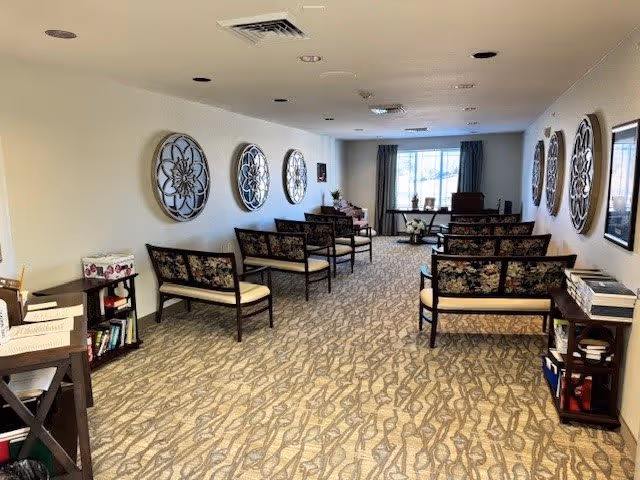 A long, narrow room with patterned carpet and floral cushioned chairs arranged in rows facing a window at the far end. The walls are decorated with large circular metal wall art and framed pictures. There are small tables and shelves with books and papers along the sides of the room.
