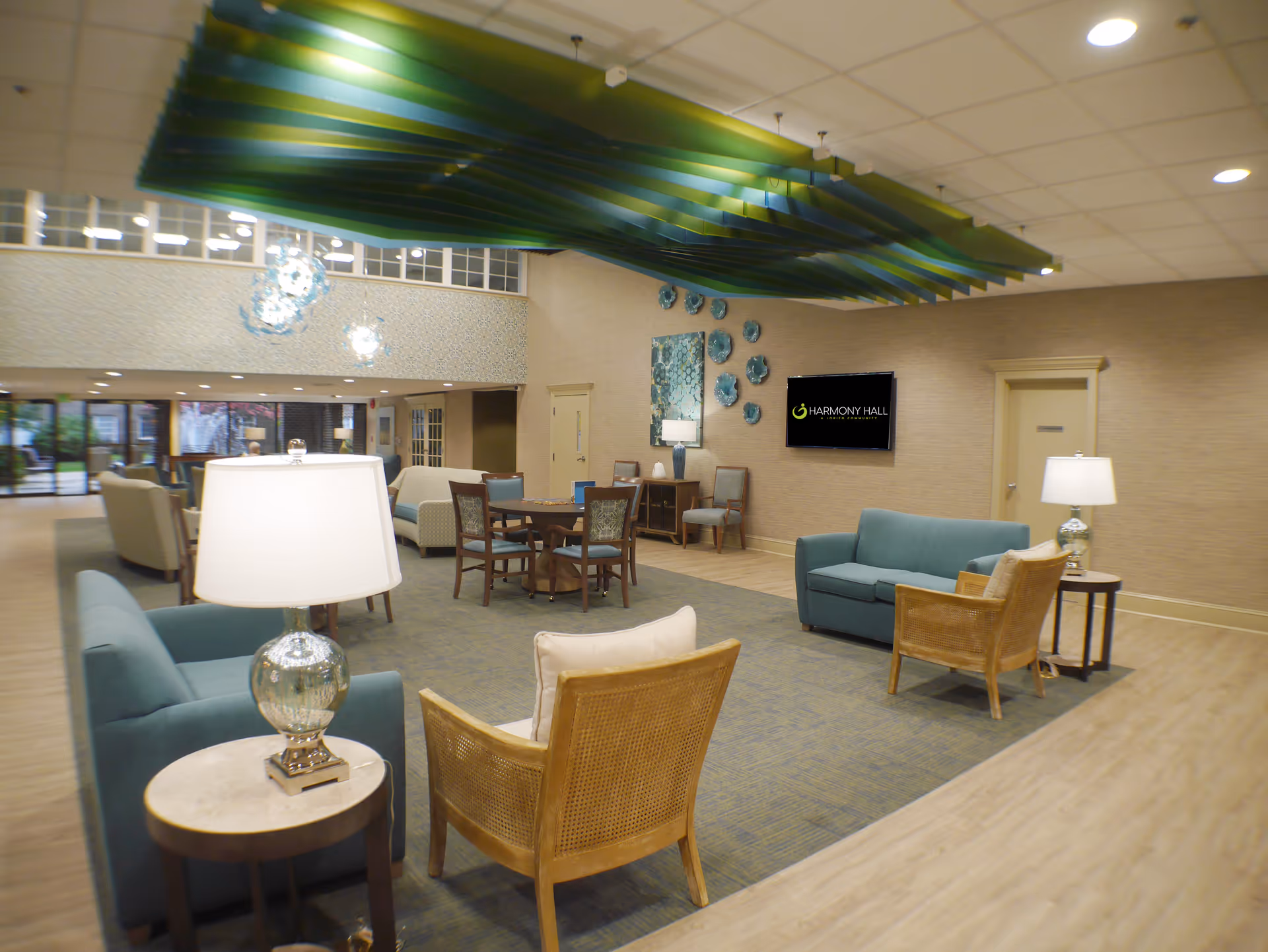 Open, well-lit lounge area with sofas, armchairs and tables under a decorative green ceiling feature and a wall-mounted TV reading 'Harmony Hall'.