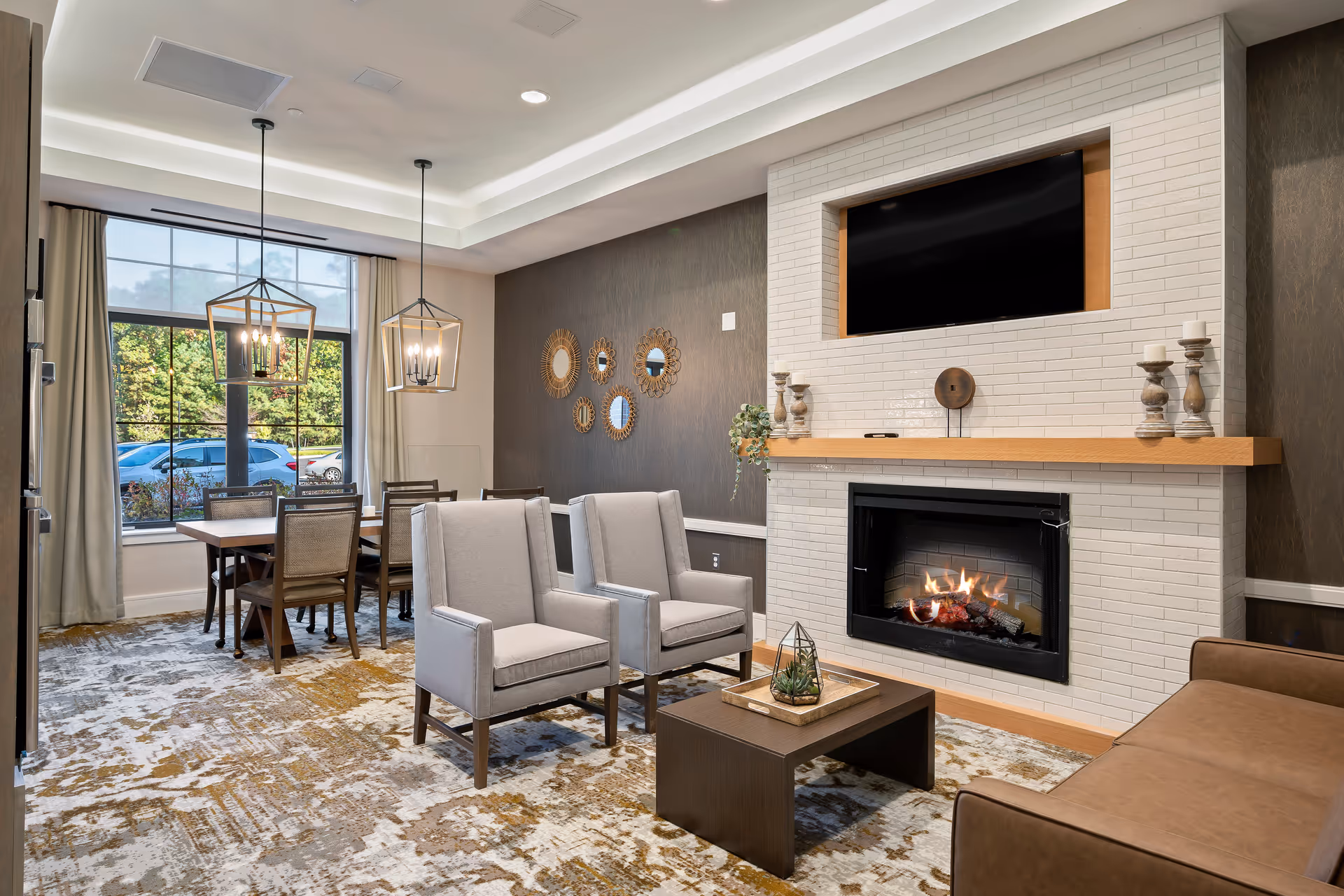 A cozy living room area with two gray armchairs and a brown sofa arranged around a dark wooden coffee table. The room features a white brick fireplace with a fire burning inside and a mounted flat-screen TV above it. On the mantel are decorative candle holders and a small plant. Behind the seating area is a dining table with six chairs, illuminated by two modern pendant lights. A large window with curtains lets in natural light and shows parked cars and greenery outside. The walls have dark textured wallpaper with decorative round mirrors.