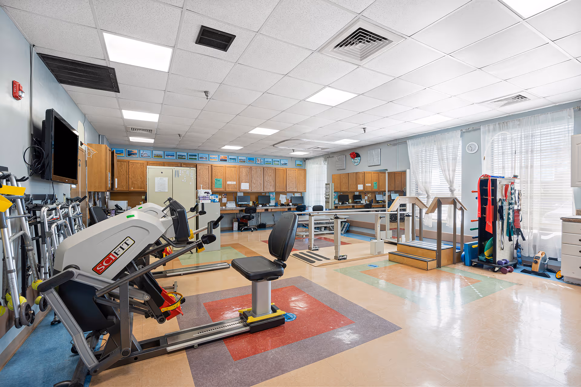 A spacious rehabilitation room with exercise equipment including a SciFit machine, parallel bars for walking practice, and various physical therapy tools. The room has a tiled floor with colorful patterns, wooden cabinets along the back wall, a wall-mounted TV, and large windows with sheer curtains allowing natural light to fill the space.