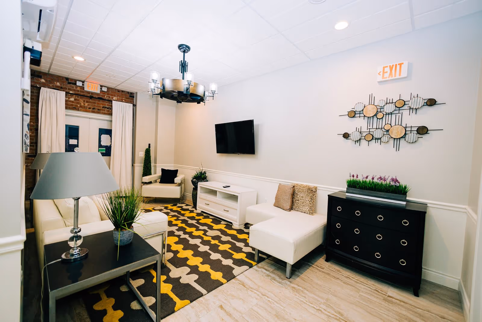A modern living room with white walls and a patterned rug in shades of yellow, gray, and black. The room features white seating including a sofa and an ottoman, a black side table with a lamp and a plant, a wall-mounted flat-screen TV, a black chest of drawers with a planter of purple flowers on top, and a decorative metal wall art piece. There is an exit sign above a door with white curtains and exposed brick around the door frame.