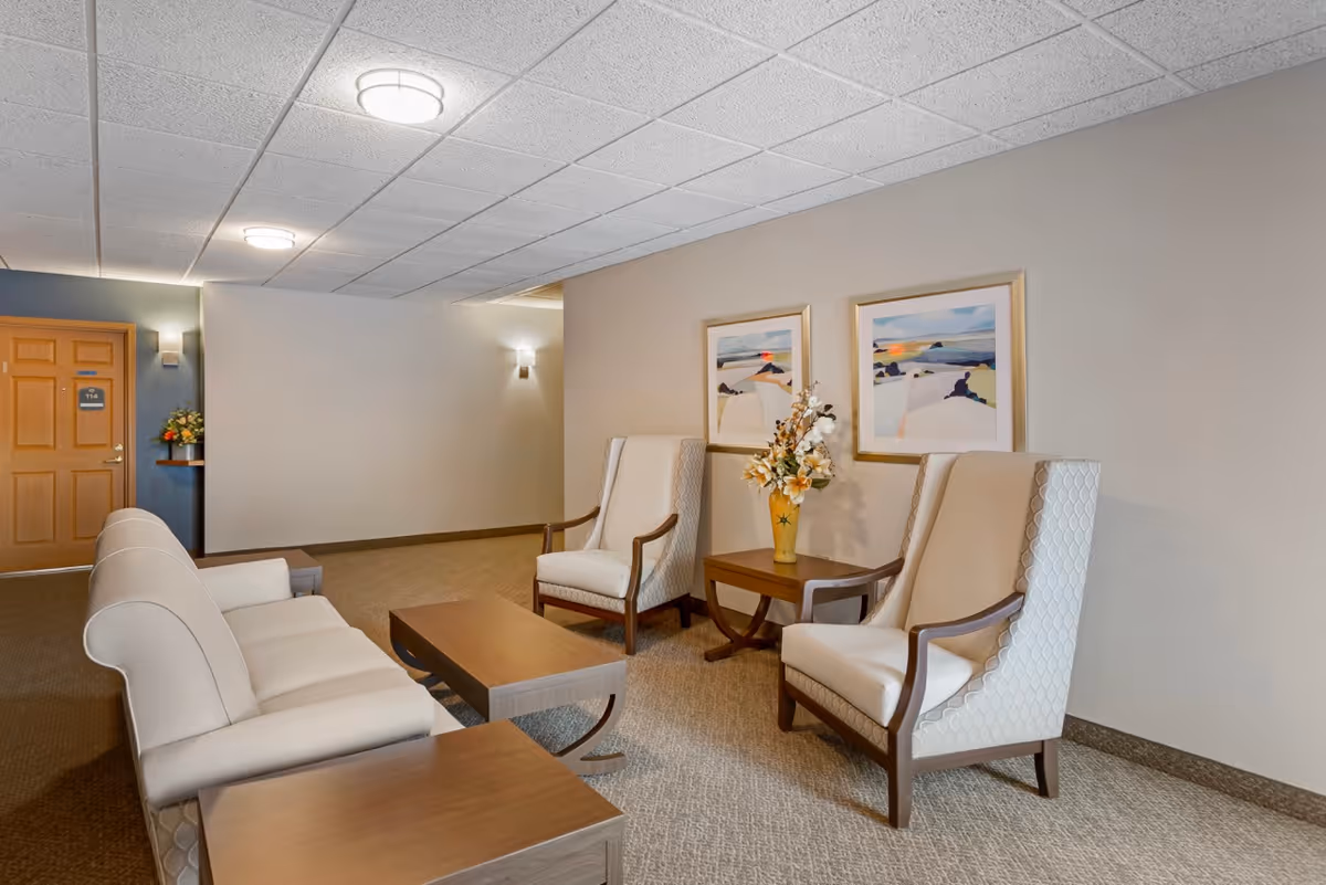 A cozy senior living facility lounge area with a beige sofa, two beige armchairs, a wooden coffee table, and a side table with a vase of flowers. Two framed landscape paintings hang on the wall behind the armchairs. The room has a carpeted floor and a ceiling with recessed lighting. A wooden door with the number 114 is visible in the background.