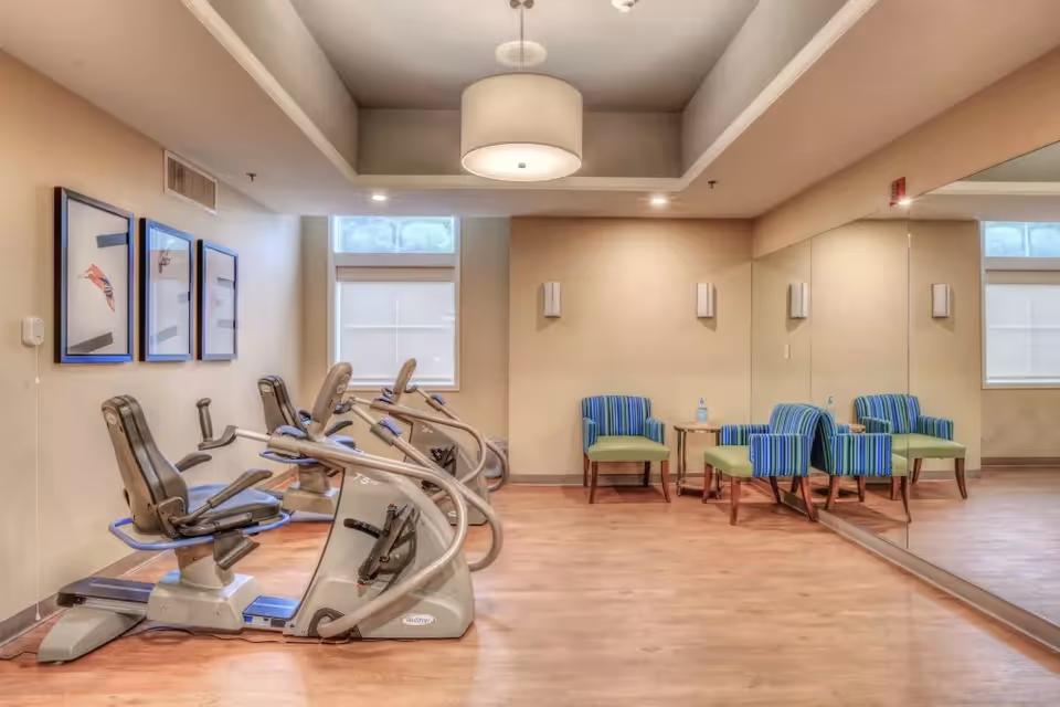 A small exercise room with three stationary recumbent bikes lined up on the left side. On the right side, there are two blue and green striped chairs with a small wooden table between them, placed in front of a large wall mirror. The room has light beige walls, wooden flooring, and a ceiling light fixture.