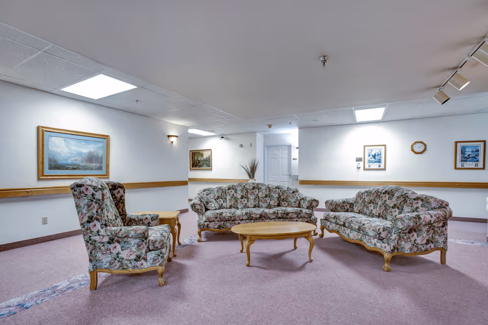 A senior living facility common area with floral upholstered sofas and armchairs arranged around a wooden coffee table. The room has light purple carpet, white walls with wooden chair rails, framed paintings, a clock, and overhead fluorescent lighting.