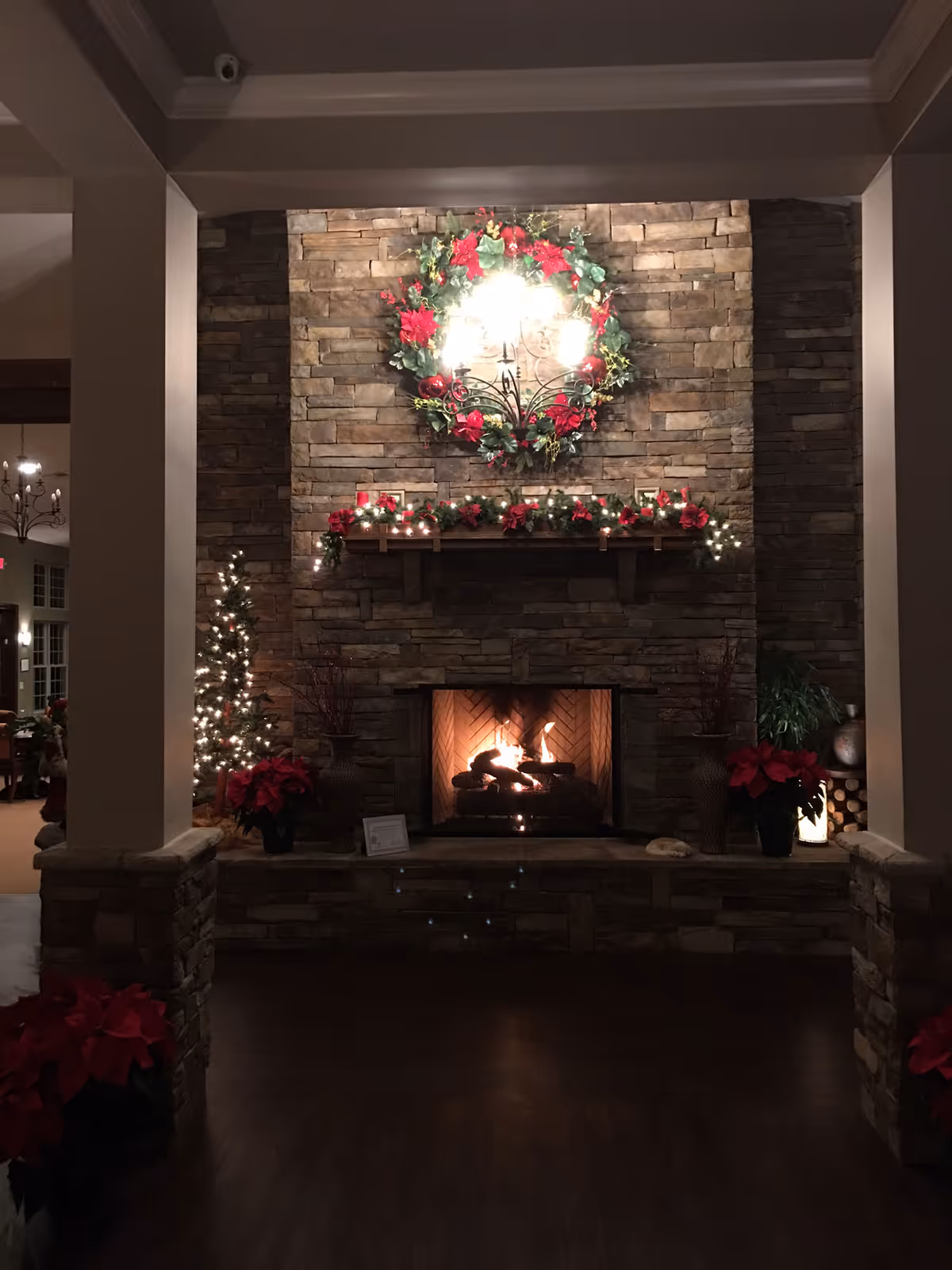 A cozy stone fireplace decorated for the holidays with a large wreath adorned with red flowers and greenery above the mantel. The mantel is decorated with garland and small lights. There are potted poinsettia plants on either side of the fireplace and a small lit Christmas tree to the left. The room has a warm, inviting ambiance with dark wood flooring and stone pillars framing the fireplace.