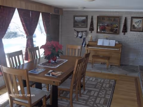 A cozy interior room with a wooden dining table and six chairs. The table has a red poinsettia plant and some magazines on it. Behind the table, there is a piano with sheet music and a bench. The walls are decorated with framed pictures and two small Christmas trees. Large windows with red curtains let in natural light, and a patterned rug covers part of the wooden floor.