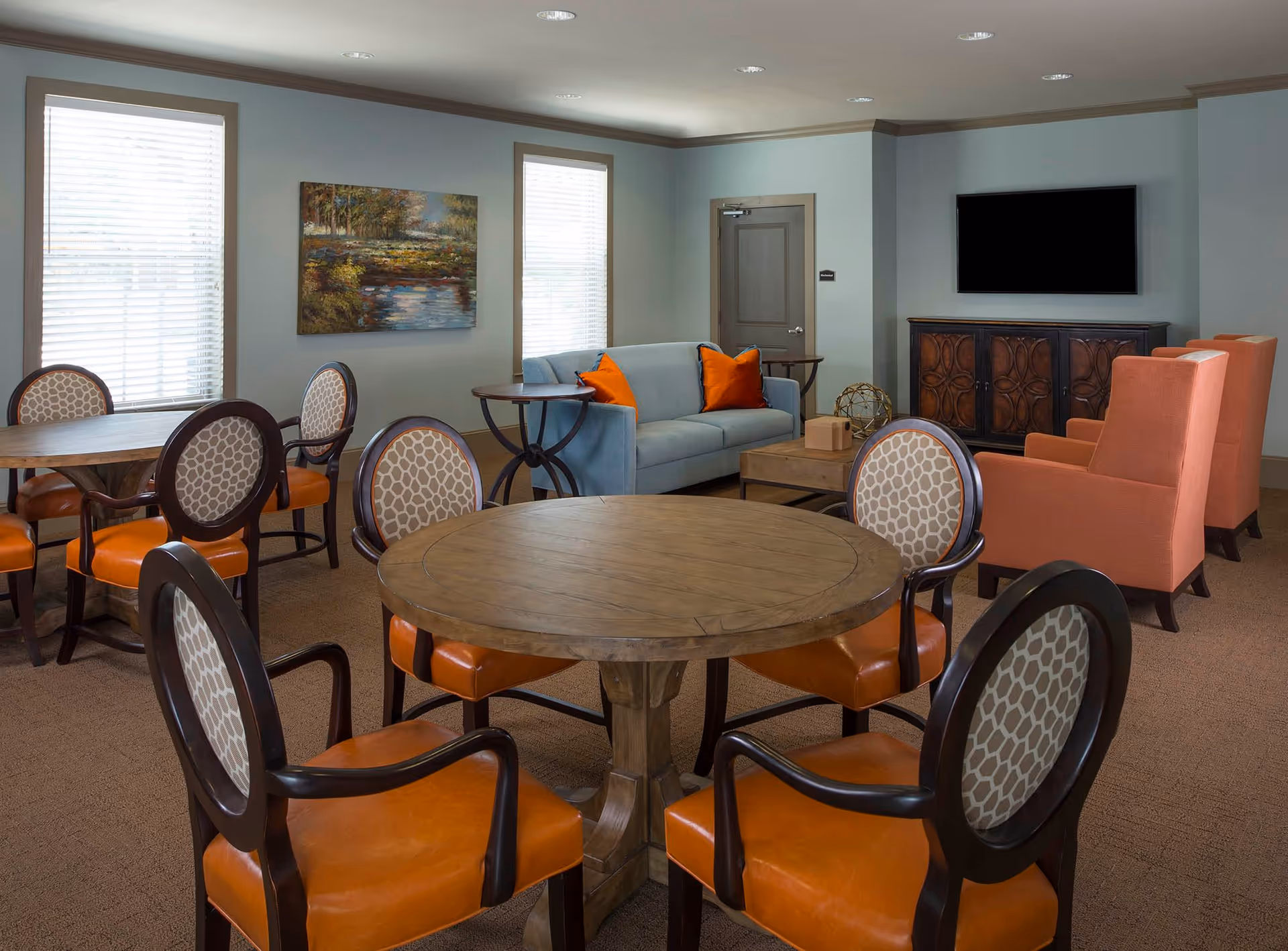 A cozy common area in an assisted living facility featuring round wooden tables with cushioned chairs that have orange seats and patterned backs. In the background, there is a light blue sofa with orange pillows, two orange armchairs, a wooden coffee table, a TV mounted on the wall above a decorative wooden cabinet, and two windows with blinds. A landscape painting hangs between the windows.