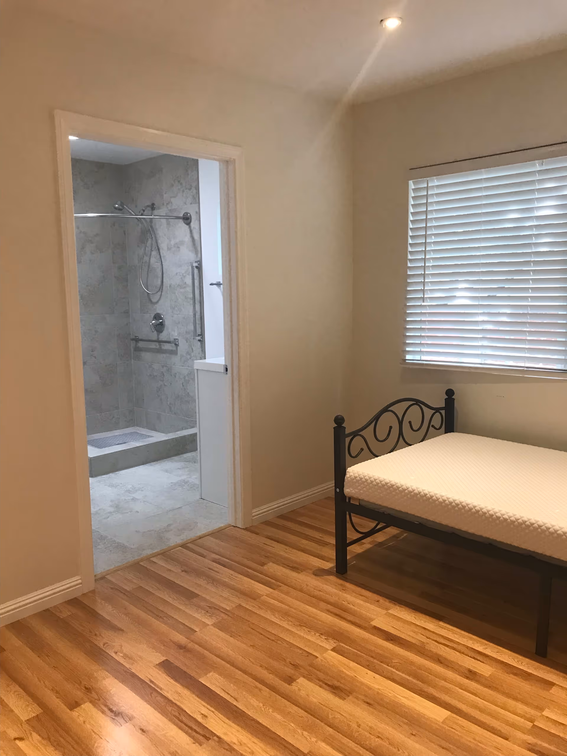 A simple bedroom with wooden flooring, a metal-framed bed with a mattress, a window with closed blinds, and an open doorway leading to a tiled bathroom with a walk-in shower and grab bars.
