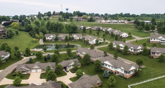 Aerial view of a retirement community with multiple single-story and two-story houses surrounded by green lawns, trees, and paved roads. There is a small pond in the center and a water tower visible in the distance under a cloudy sky.
